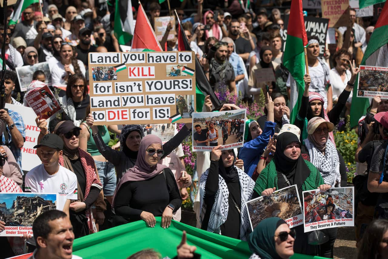A group of protesters holding placards in support of Palestinians. Some are wearing Palestinian keffiyeh and others holding Palestinian flags.