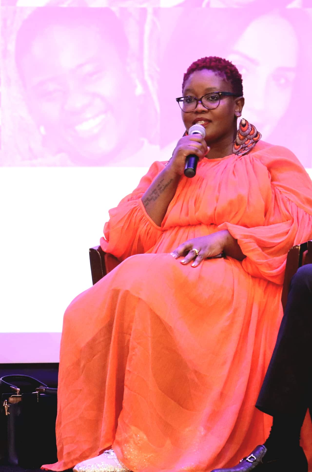 An African woman wearing an orange dress speaks into a microphone