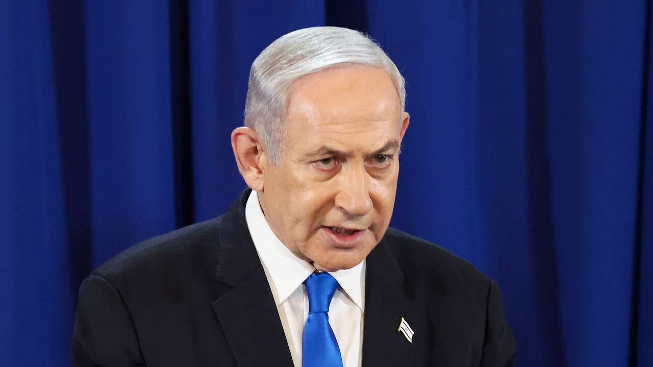 Benjamin Netanyahu wearing a suit, white shirt and blue tie. He is standing in front of a blue curtain.