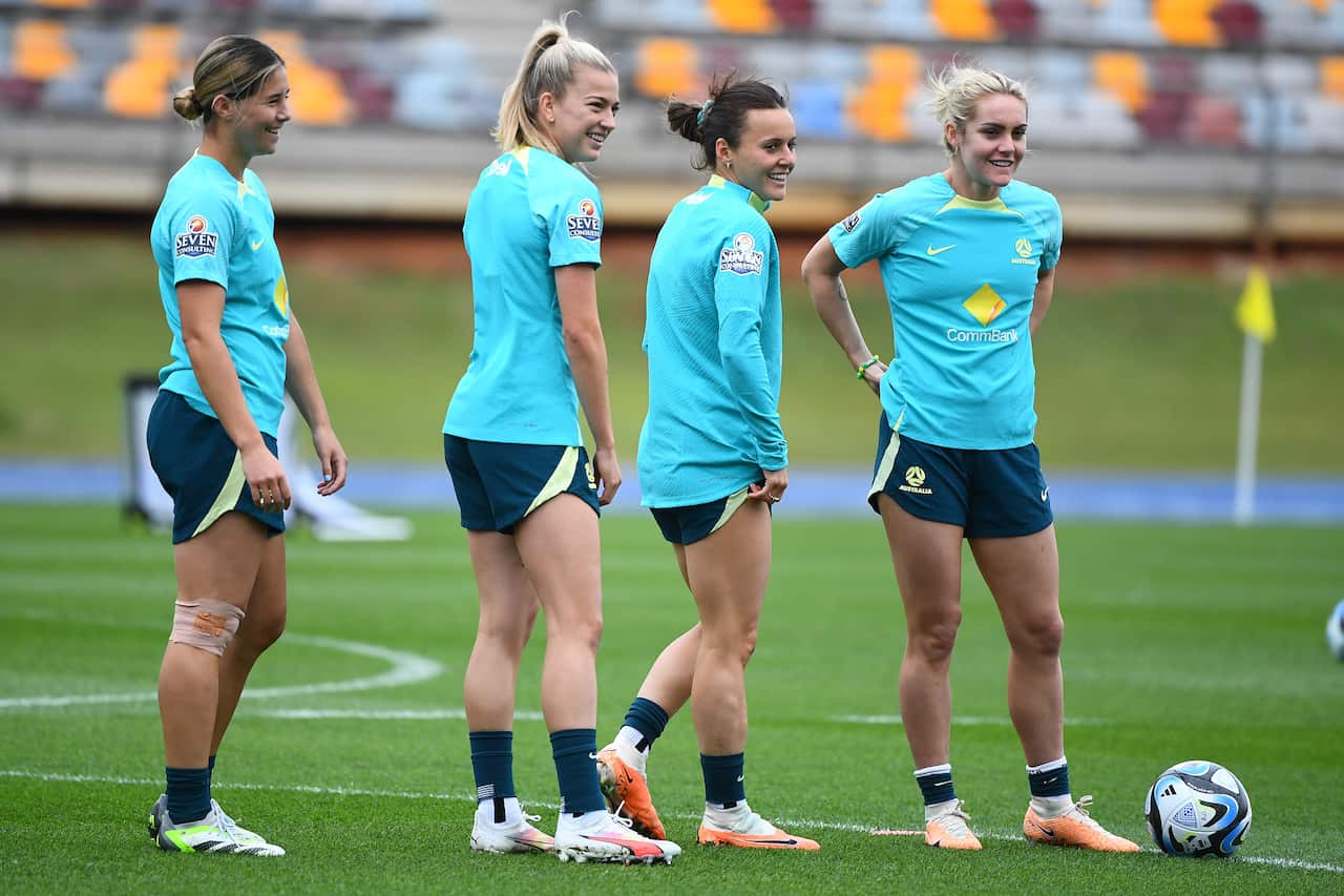 Four women footballers, wearing aqua-coloured shirts and dark green shorts standing on a grass field at training