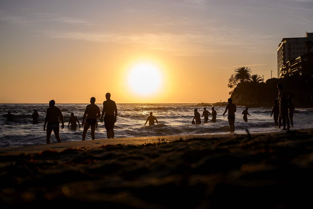People entering the water at a beach, with a bright sun shining above them.