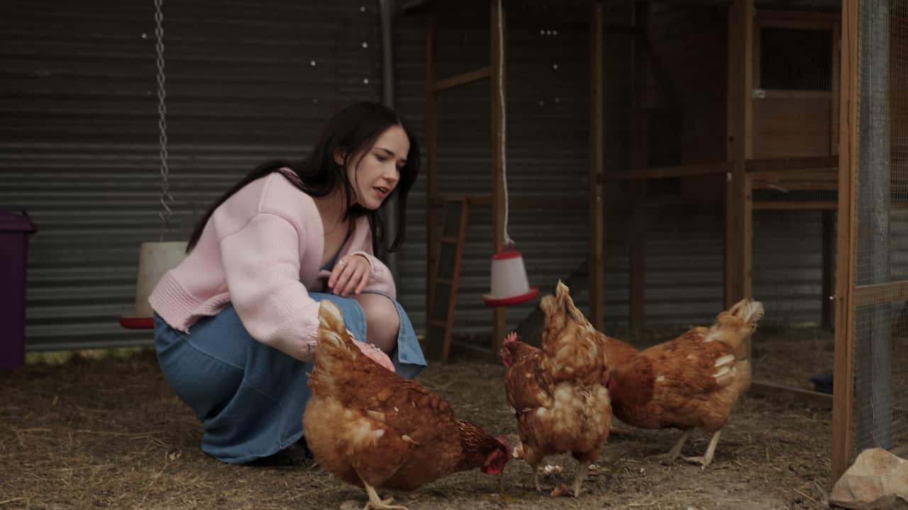 A young woman crouches beside three chickens who are pecking at the ground
