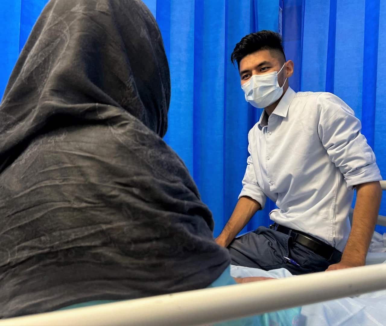 Kumail Jaffry wearing a mask and sitting on a bed. He is facing a patient whose back is turned to the camera.