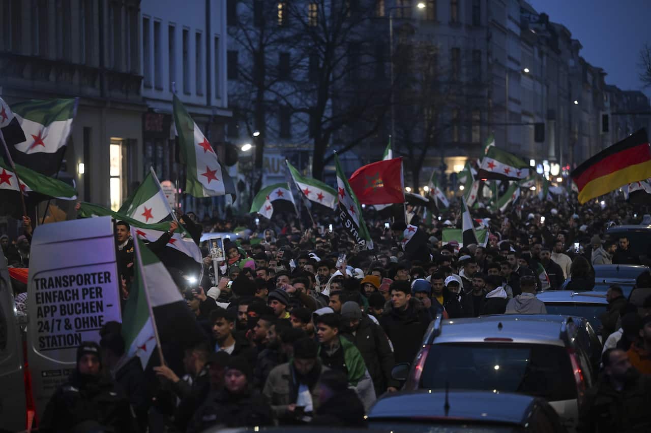 People carrying flags and posters gathered in a square, with cars visible adjacent to them.