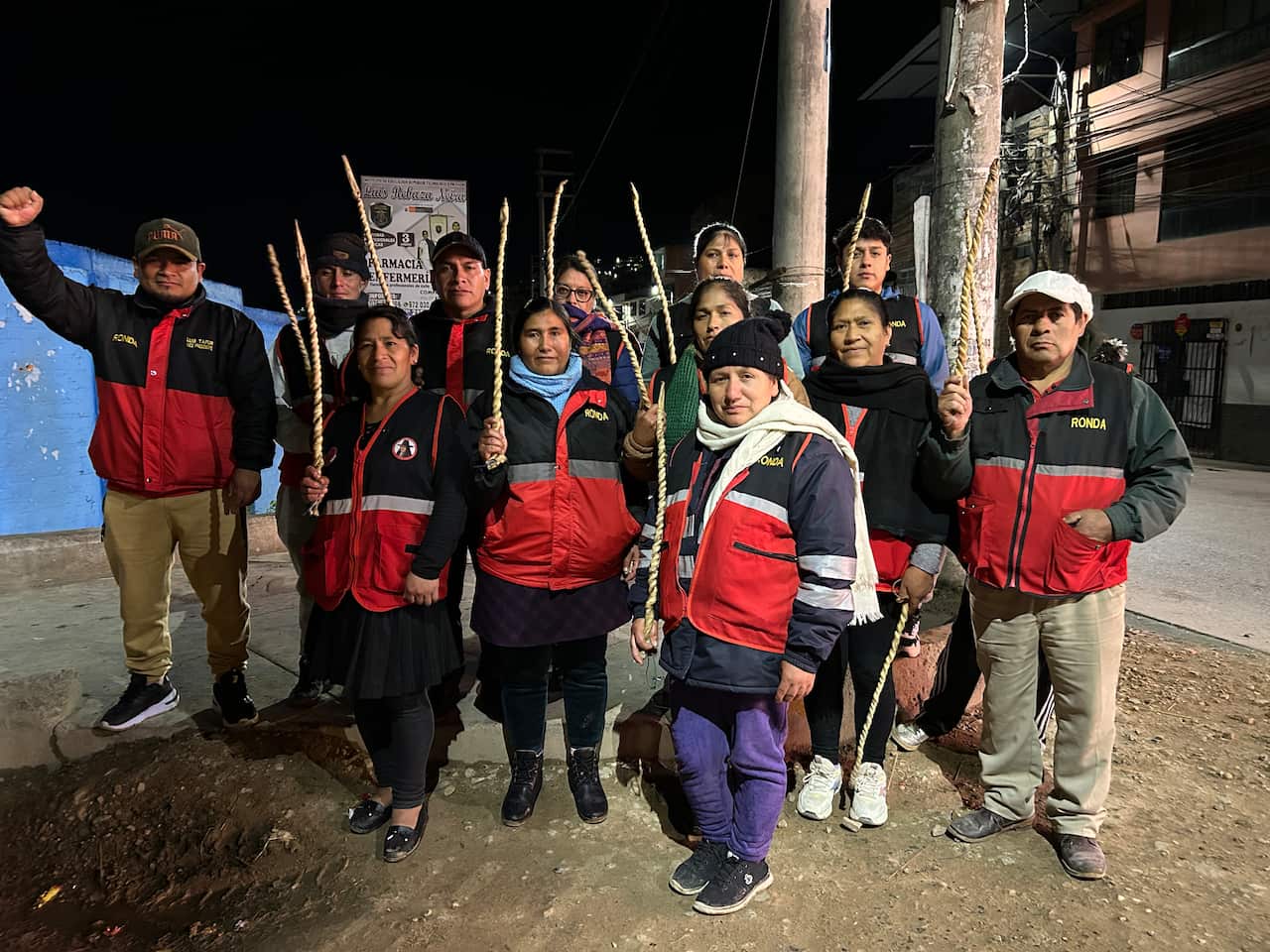 A group of men and women in matching black and red vests hold up long braided sticks. 