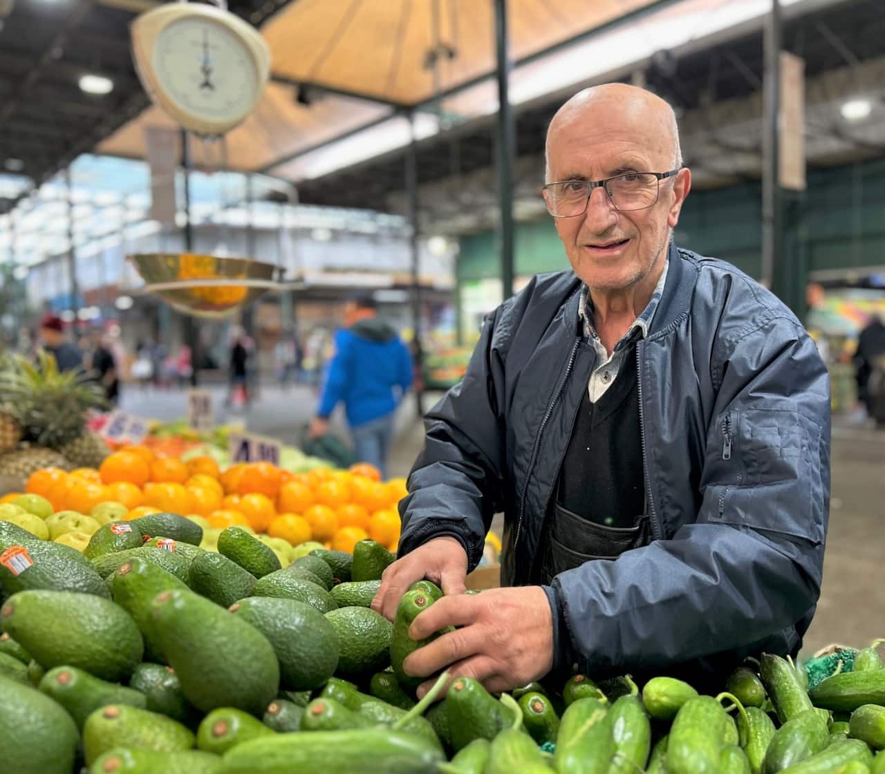  A man in a blue jacket stands holding avocados on a table in a market store.