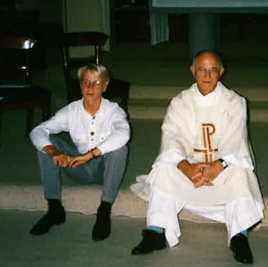 A man and a Catholic priest sit on some steps with neutral expressions on their faces.