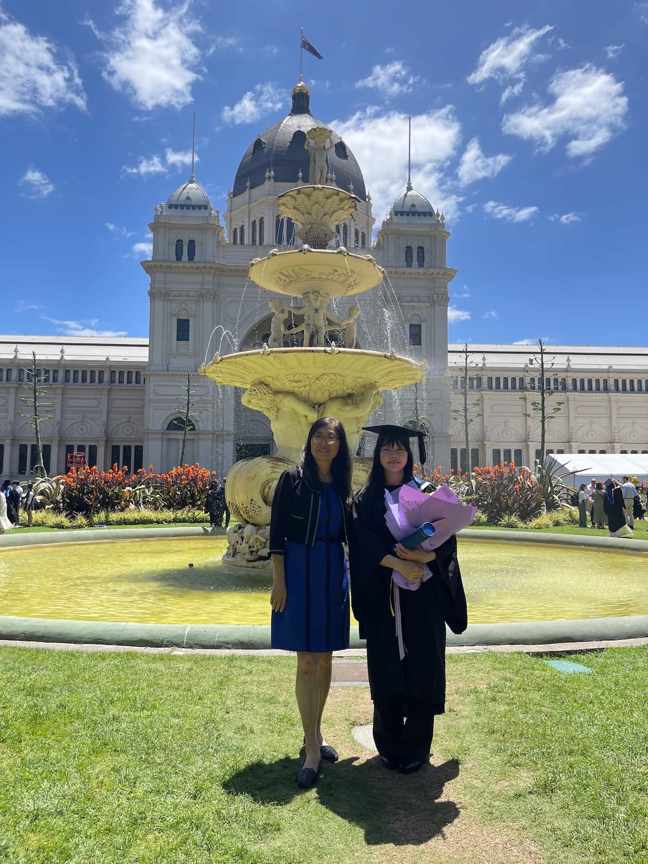A young Chinese woman in academic regalia holds a bunch of flowers, standing next to her mother in front of the Royal Exhibition Building in Melbourne. 