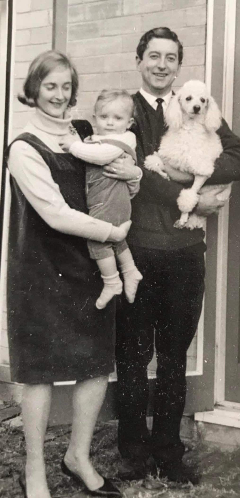 A black and white photo of a mum and dad standing together and smiling as they hold their baby daughter and a dog.