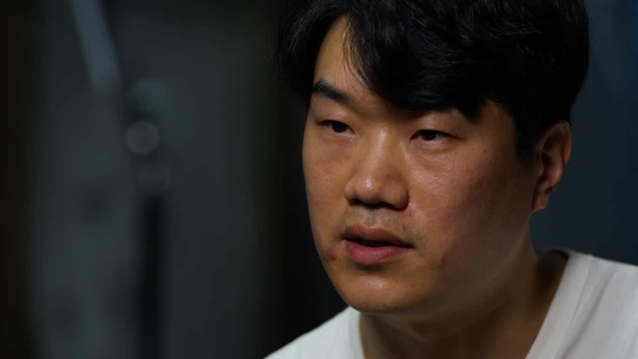 A young man wearing a white T shirt looks at the camera with a serious expression on his face.
