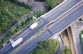 Three cars and two trucks parked on a two-way, four lane highway. There is a large crack in the middle of the highway, where the road has been damaged.