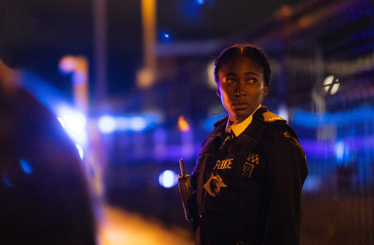 A wound woman in a police uniform stands in a night-lit street. 