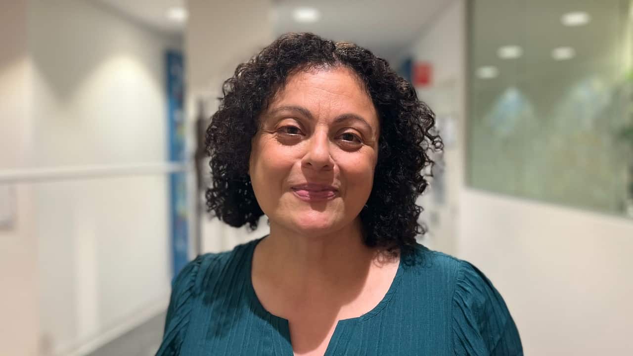 a closeup shot of a middle aged woman with curly hair in a turquoise top standing in front of a white hallway