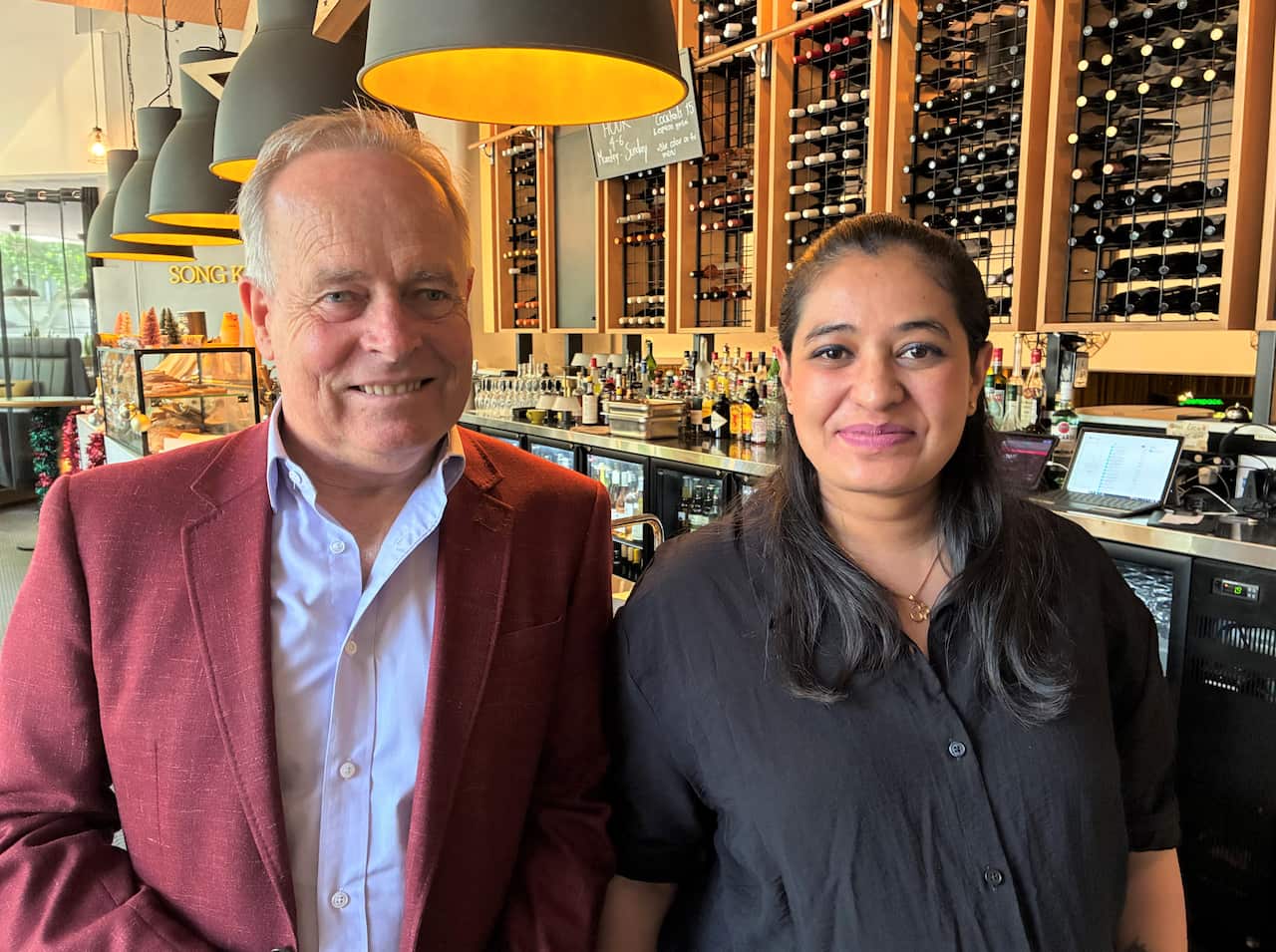 A man in a burgundy jacket stands next to a woman in a dark blue shirt with long brown hair.