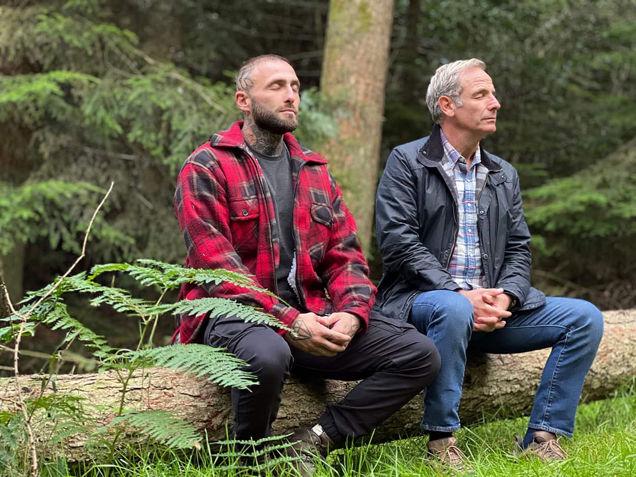 Two men sit on a tree log in the forest with their eyes closed in meditation.jpg