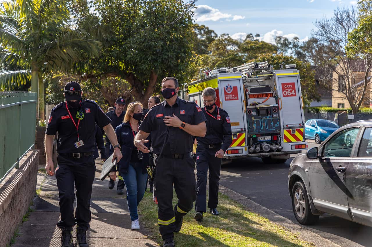 A group of firefighters walk on a suburban footpath. A fire truck is in the background