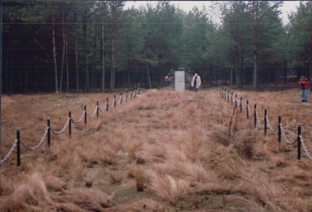A wide shot photograph of an open field with tufts of brownish grass, in front of a forest of tall, dark green trees. An area is fenced off, the spot of the mass grave, a man stands in the distance next to a headstone and a memorial site.
