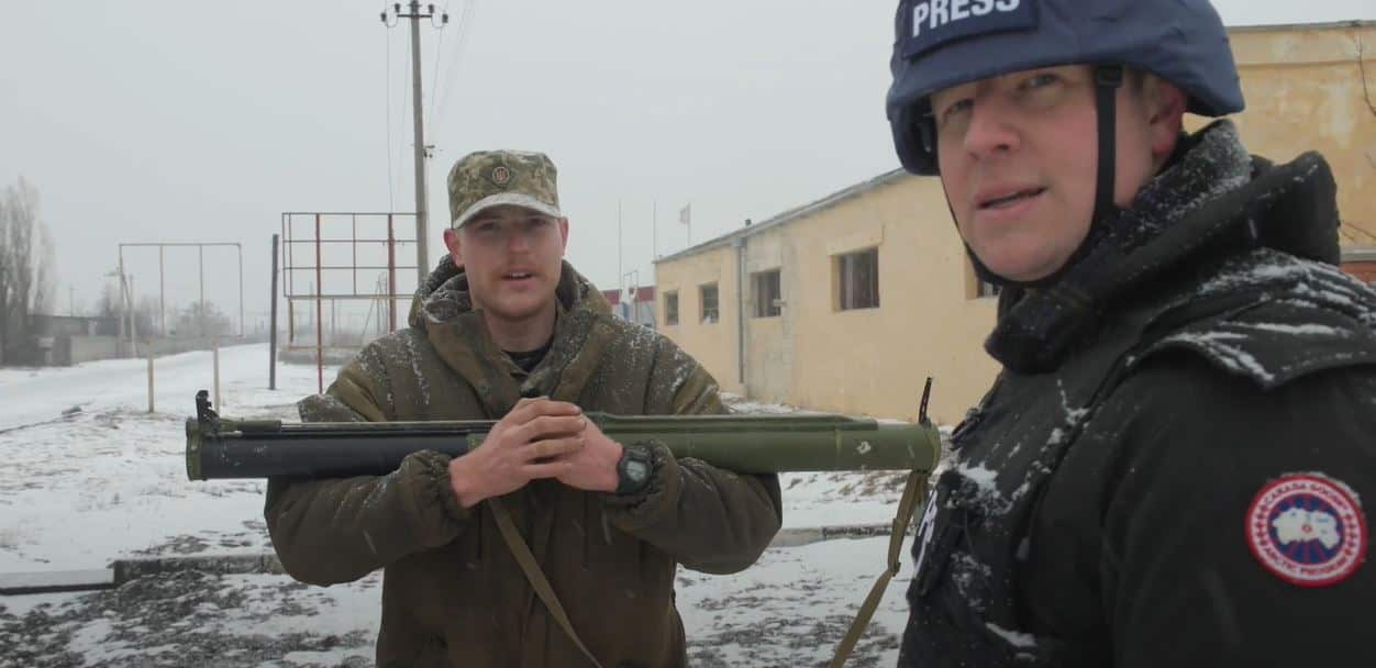 Two men standing in the snow. One man is wearing army clothes and holding a weapon across his chest. The other is wearing a dark jacket and a dark helmet with the word "Press" on it.