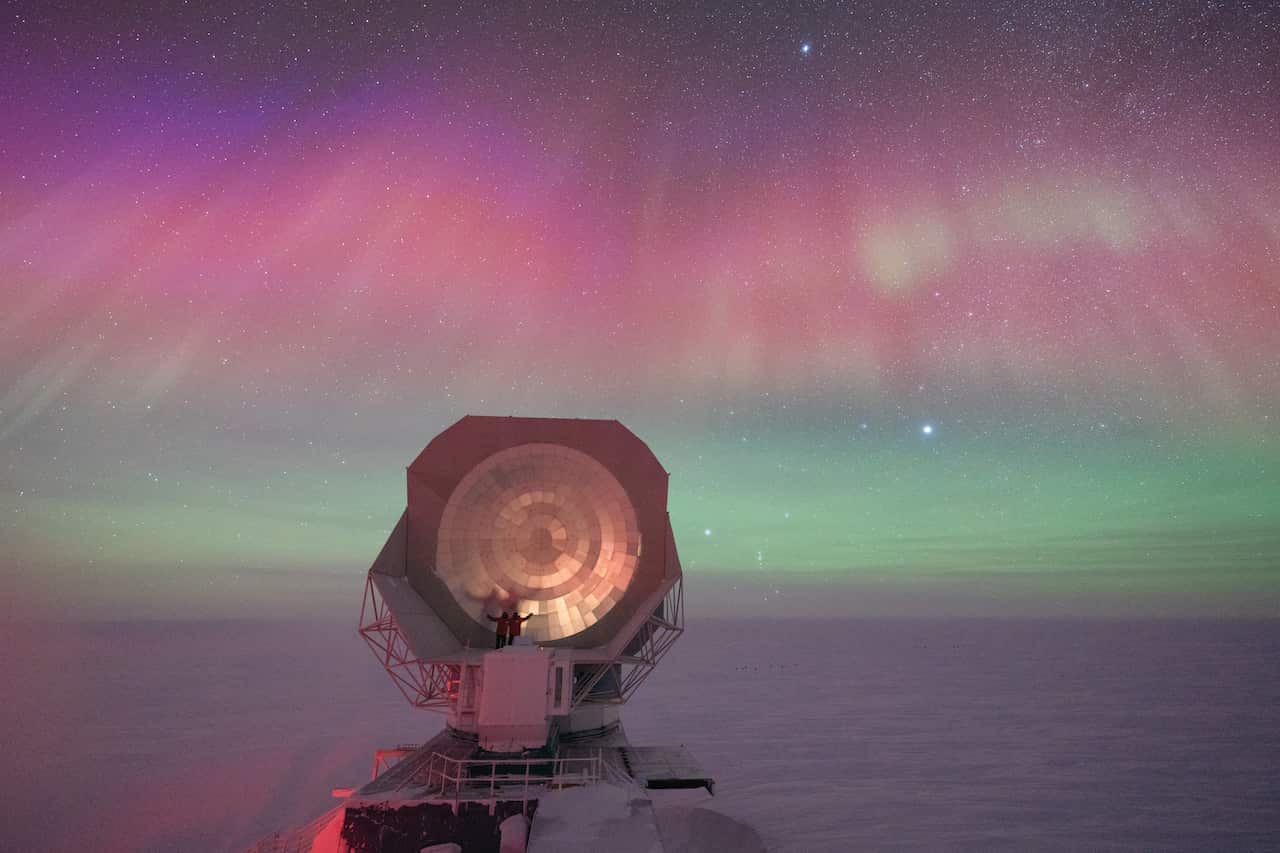Two figures stand on a large telescope with a colourful sky in the background.