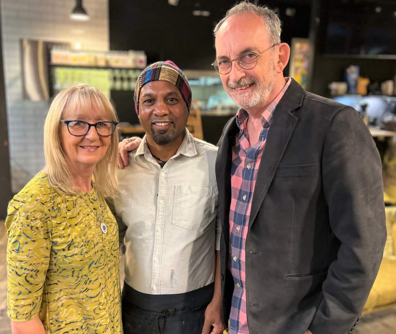 A woman in a gold dress stands next to a man in a grey shirt, and another man in a suit jacket.