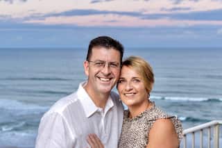 a middle aged couple pose together in front of the sea at sunset