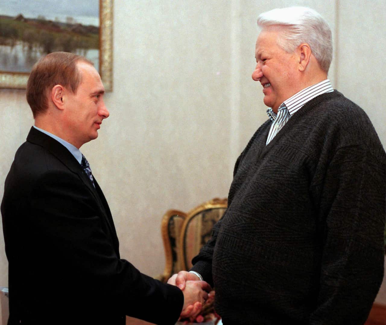 Two men are shaking hands and looking at each other. One is older, with white hair, and he is smiling.