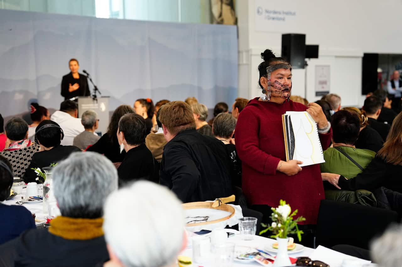 A woman in an all black outfit speaks at a podium as the audience listens. There's a woman standing amid the audience in a maroon dress holding a notepad with a sketch of a woman's body.