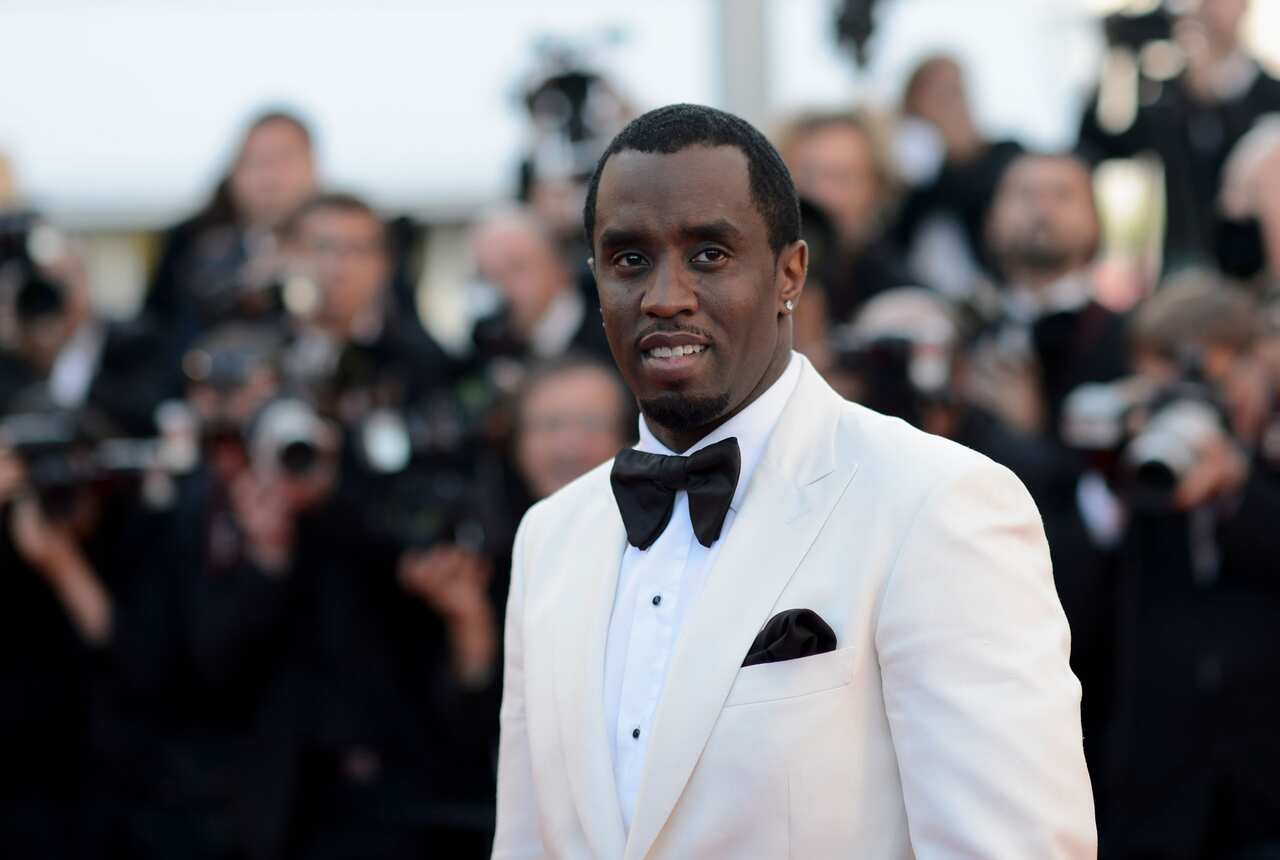 Sean "Diddy" Combs, wearing a white tuxedo, is standing in front of a large group of photographers.