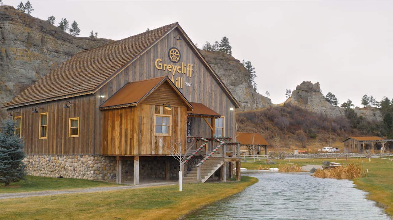 A wooden building with a sign labelled 'Greycliff Mill' sits on a green field with a river running through it. Behind the building are high rocky cliff faces. 