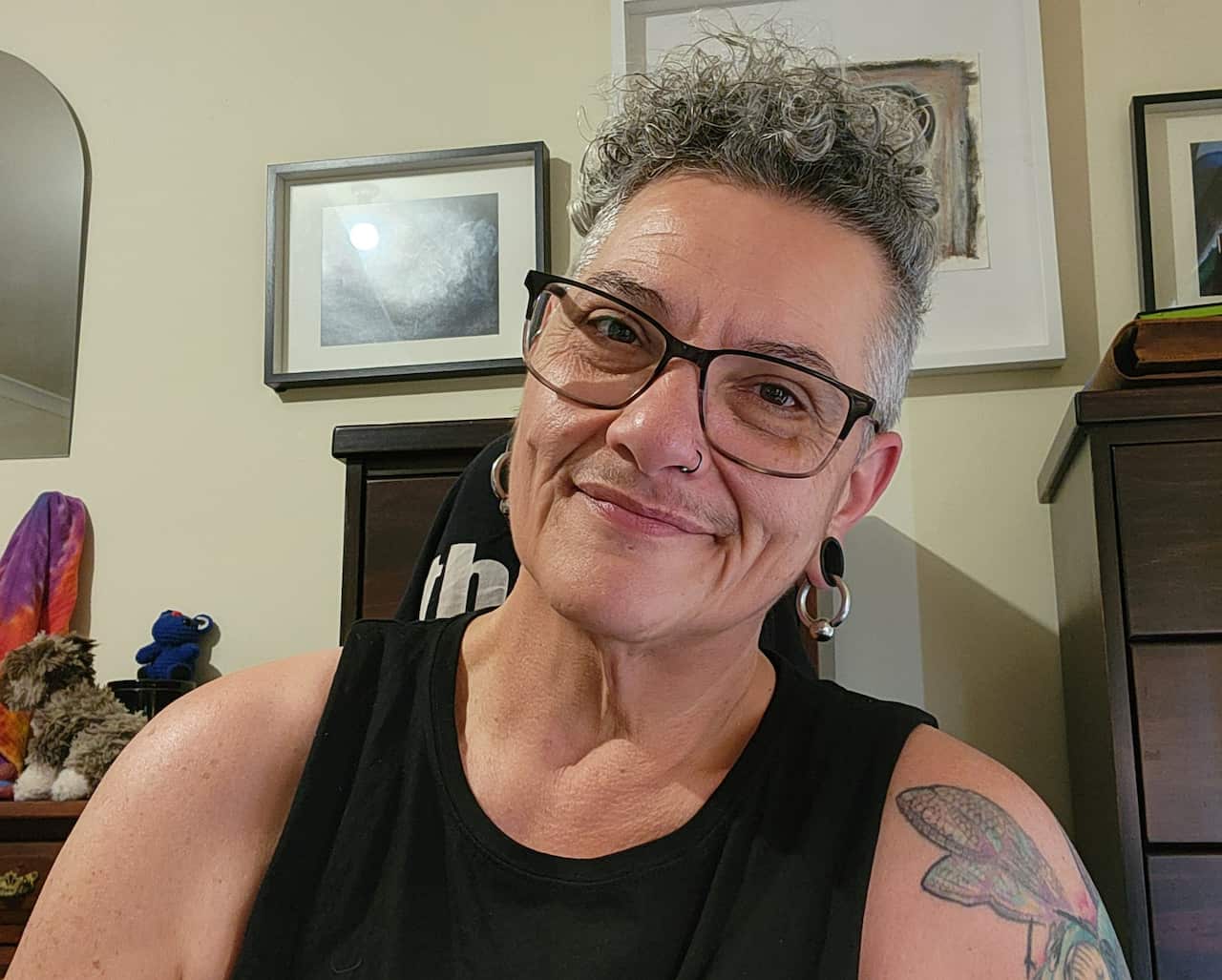 A person wearing a black singlet and glasses, smiling with their head tilted