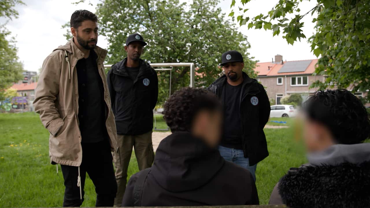 Three men stand in front of two teenagers with blurred-out faces sitting on a bench. The man on the right is wearing a beige raincoat, while two other men are wearing black parkas and black baseball caps