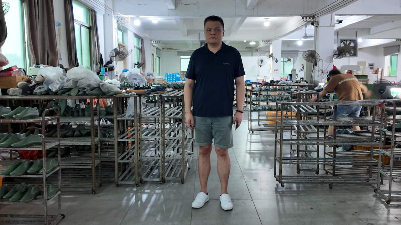 A man wearing shorts and black polo shirt stands on the floor of a clothing factory of almost-empty racks. The man is looking serious