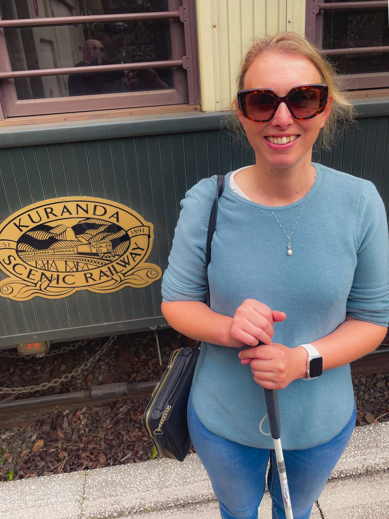 A woman wearing glasses and holding a cane in front of a train carriage that has a logo reading 'Kuranda Scenic Railway'.