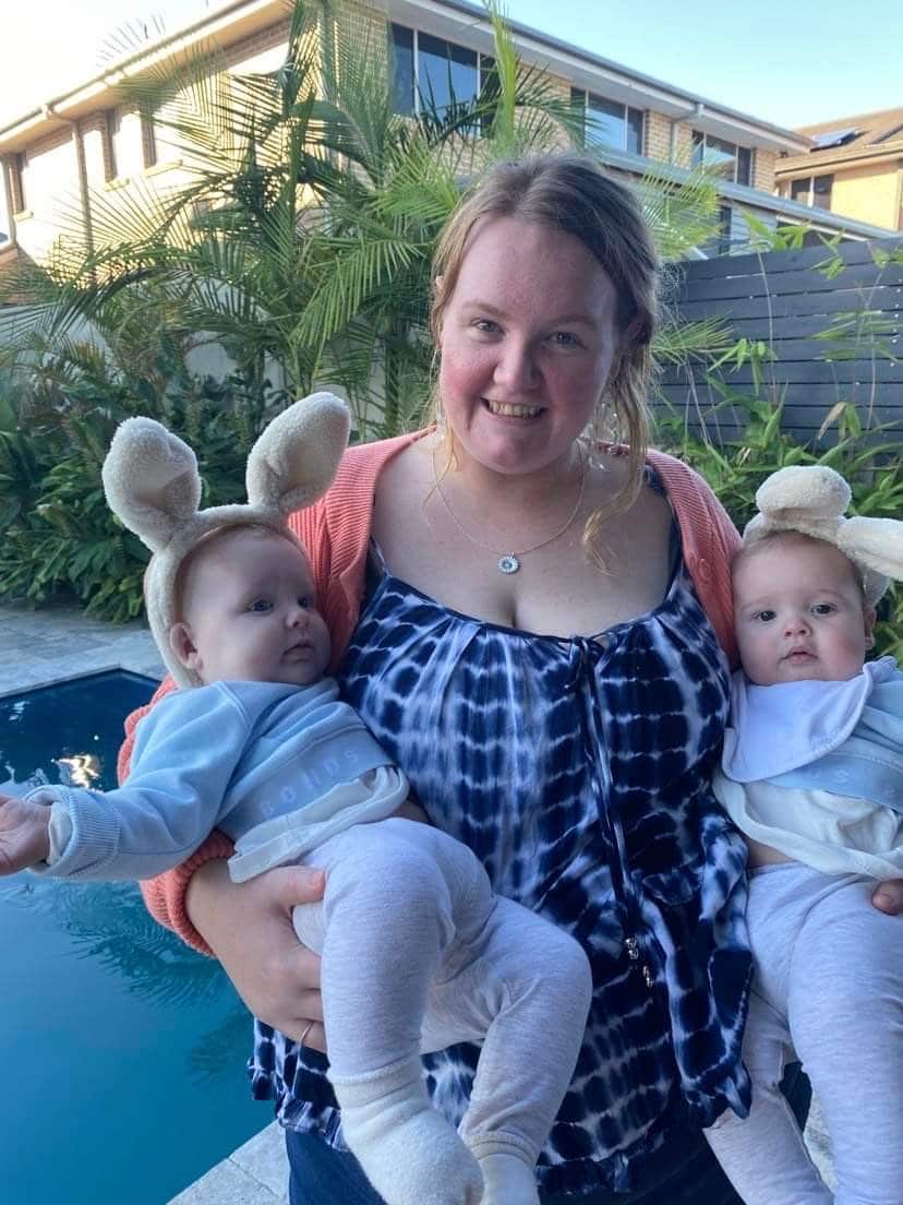 A woman stands outside next to a pool holding a pair of twin babies and smiling.