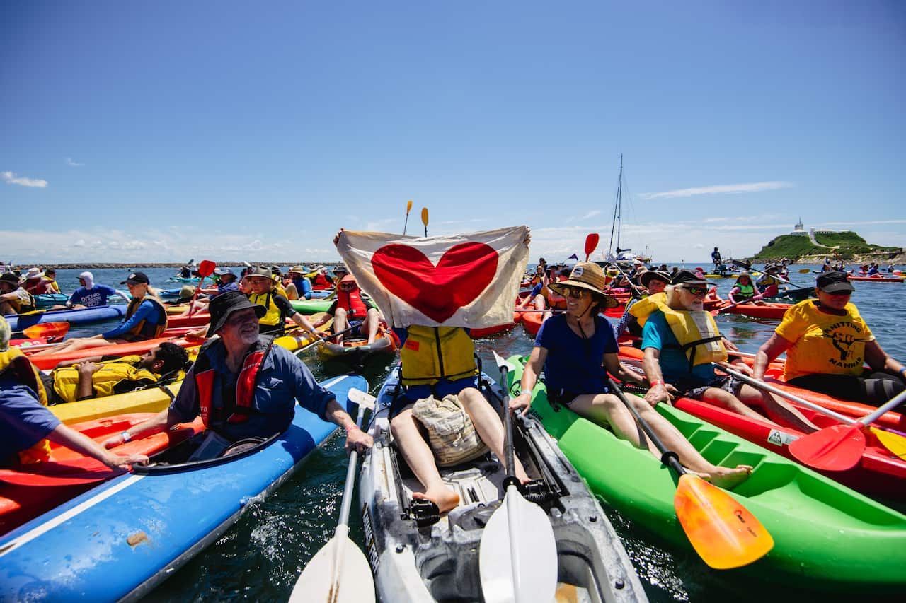 Man in a kayak holding up a banner with a red hart, while surrounded by dozens more people in kayaks.