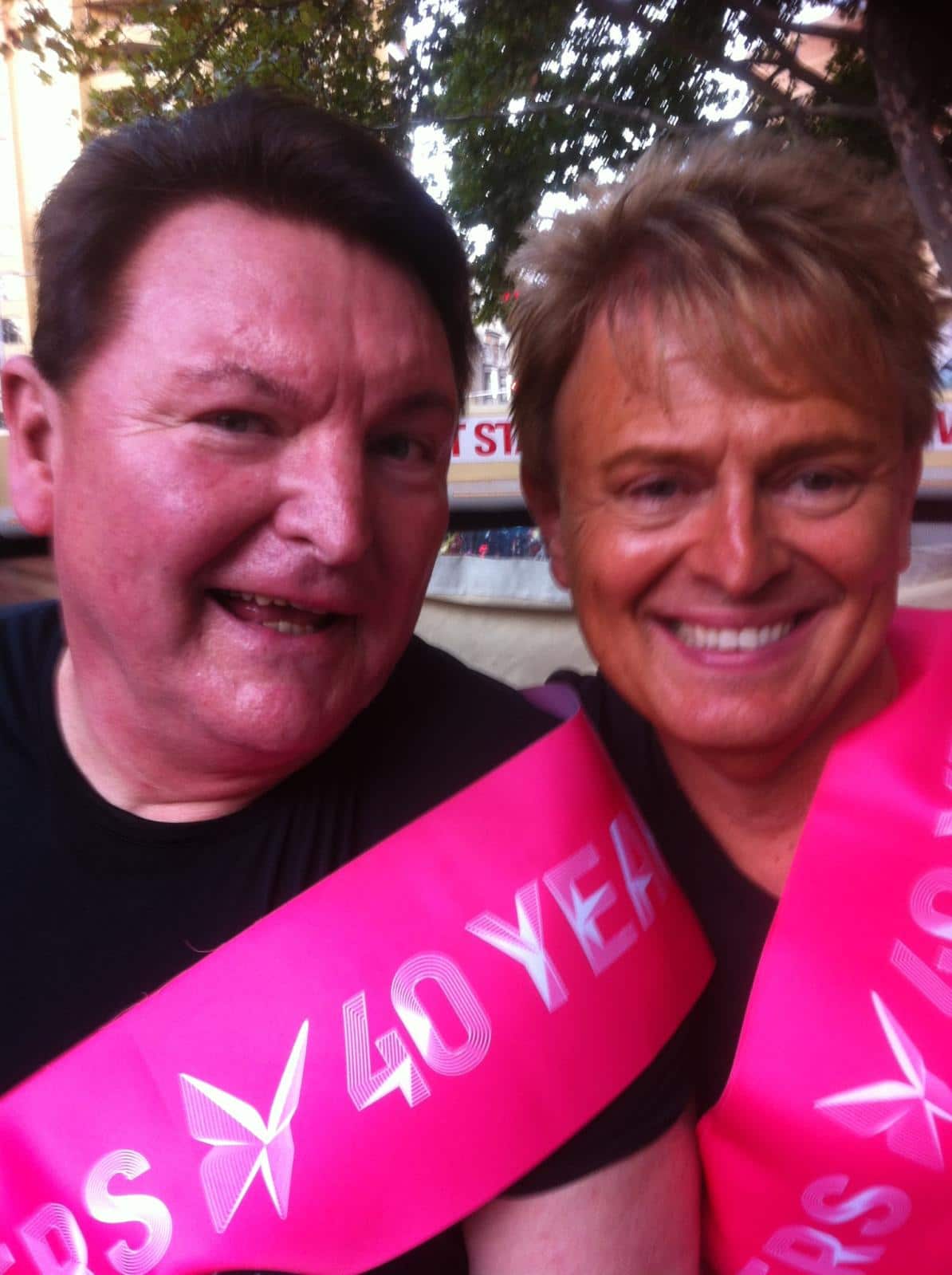 A closeup shot of two men smiling and wearing pink sashes