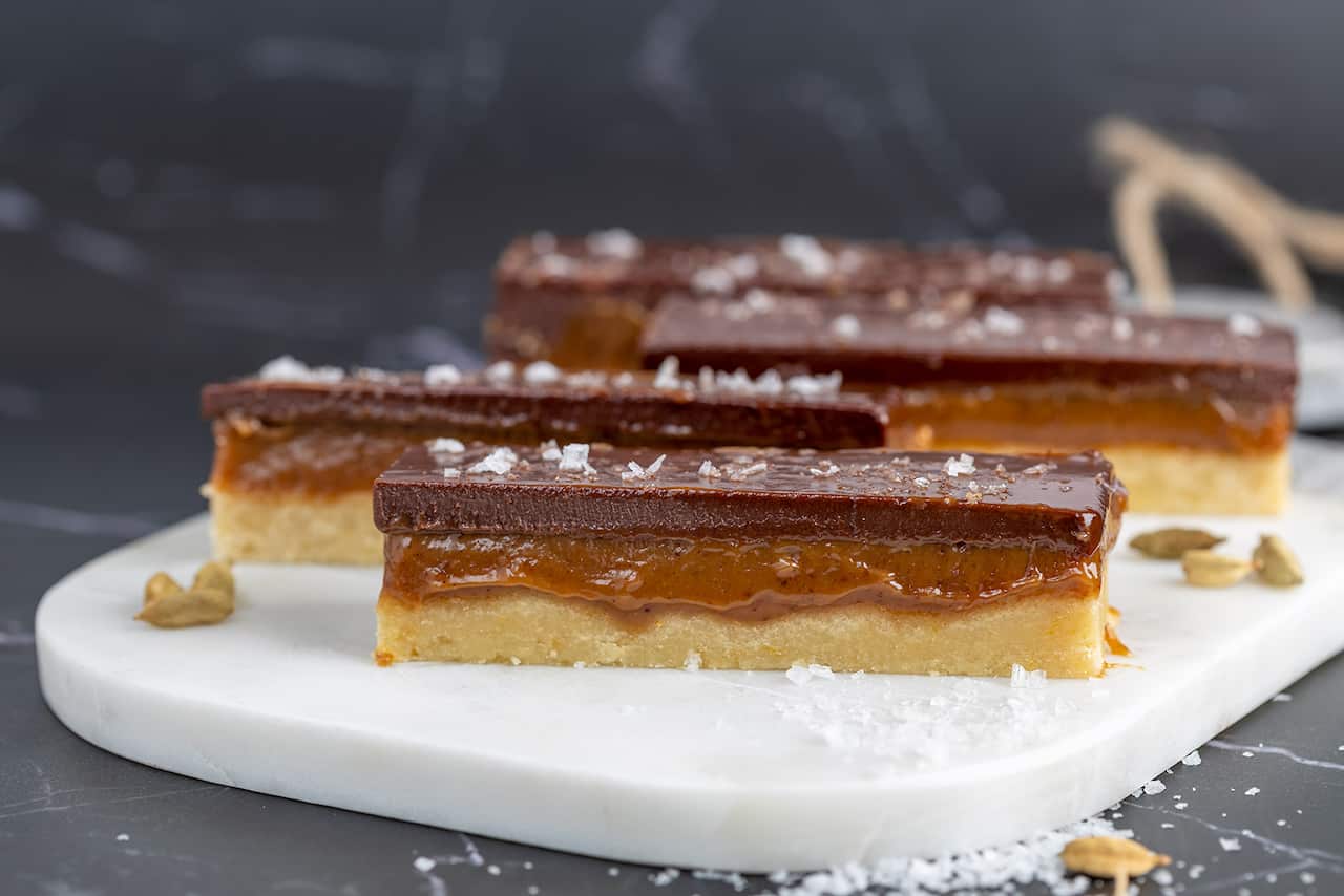 Four fingers of a rich slice sit on a white platter. The bars have three layers - a shortbread base, a gooey-looking caramel middle and a top later of chocolate, which is dusted with salt flakes. 