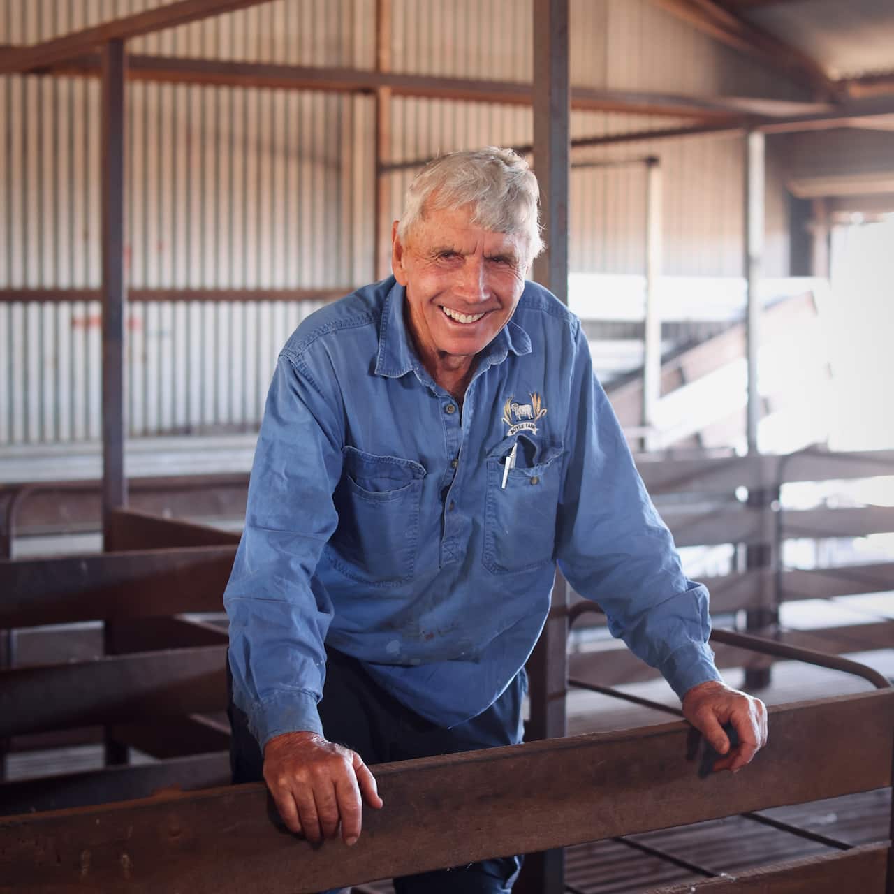 A man with short grey hair and wearing a long-sleeved blue work shirt stands smiling behind a wooden fence inside a shed