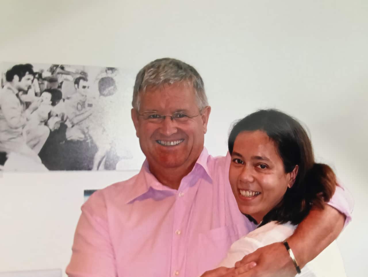 A smiling man wearing a pink shirt hugs a smiling woman wearing a white top. There is a black-and-white picture behind them of airmen feeding babies from bottles.