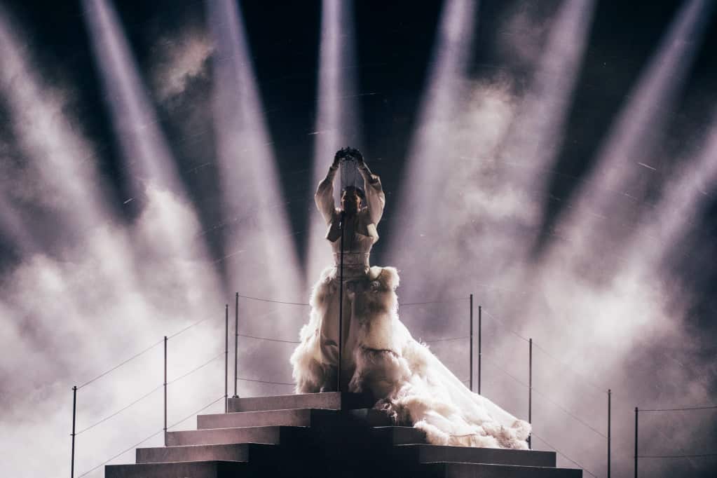 Sheldon Riley on stage wearing a white gown with his arms up, surrounded by smoke
