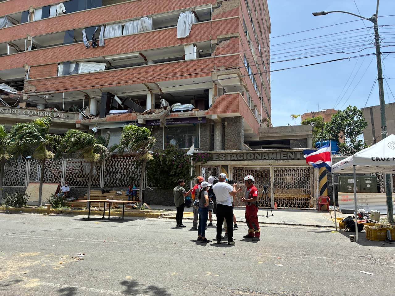 Rescuers stand in a circle on the street, with a partially demolished building behind them.