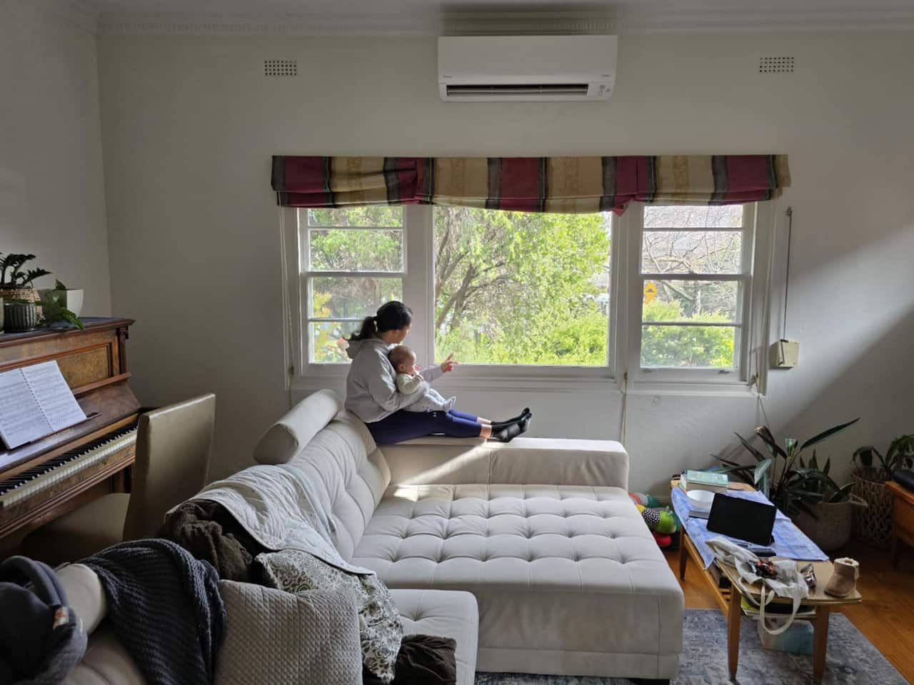 A woman sits on a sofa while holding a baby and staring out of her windows