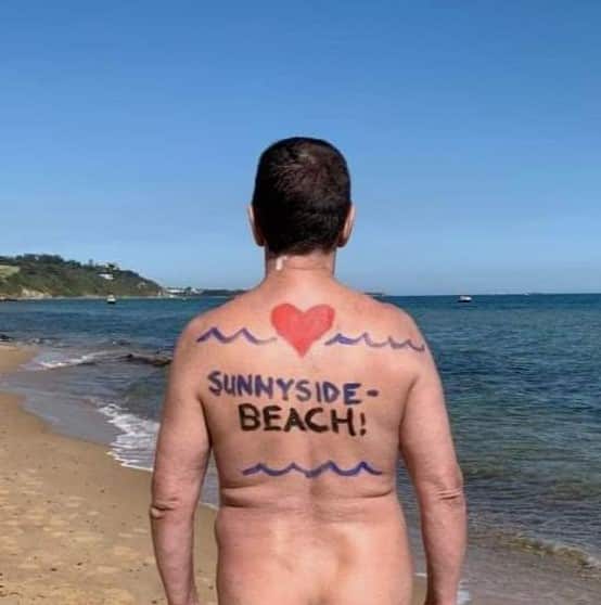 A nude man stands on a beach. Waves and a love heart are drawn on his back, along with the words "Sunnyside Beach!".