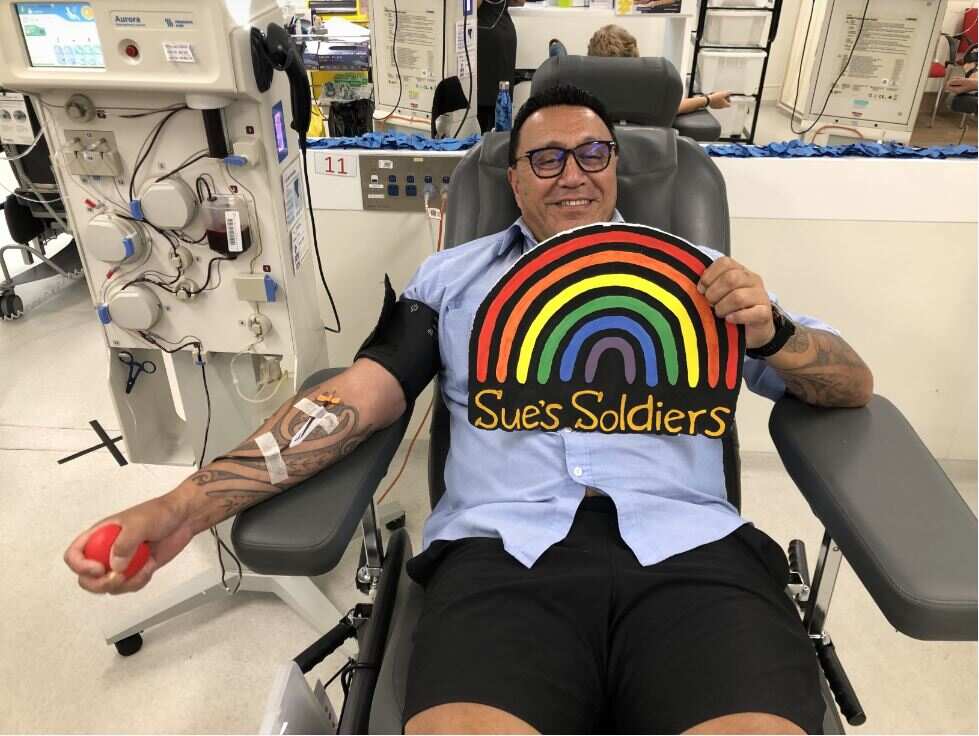 A man wearing a light blue shirt and dark blue shorts reclines in a chair as he donates blood. He he holding a sign with his left hand that features the colours of the rainbow and the words Sue's Soldiers.
