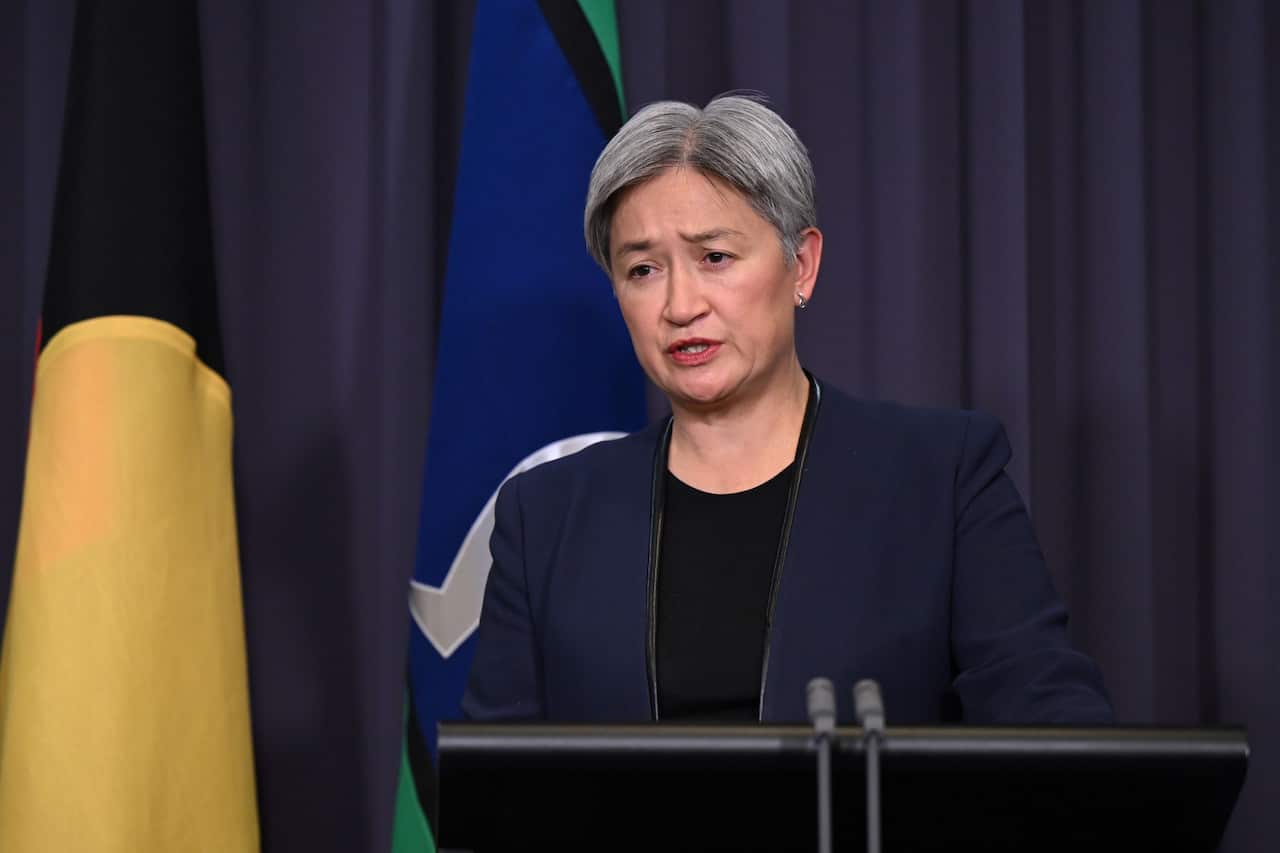 A woman with short, grey hair is speaking at a podium in front of a blue curtain and flags.