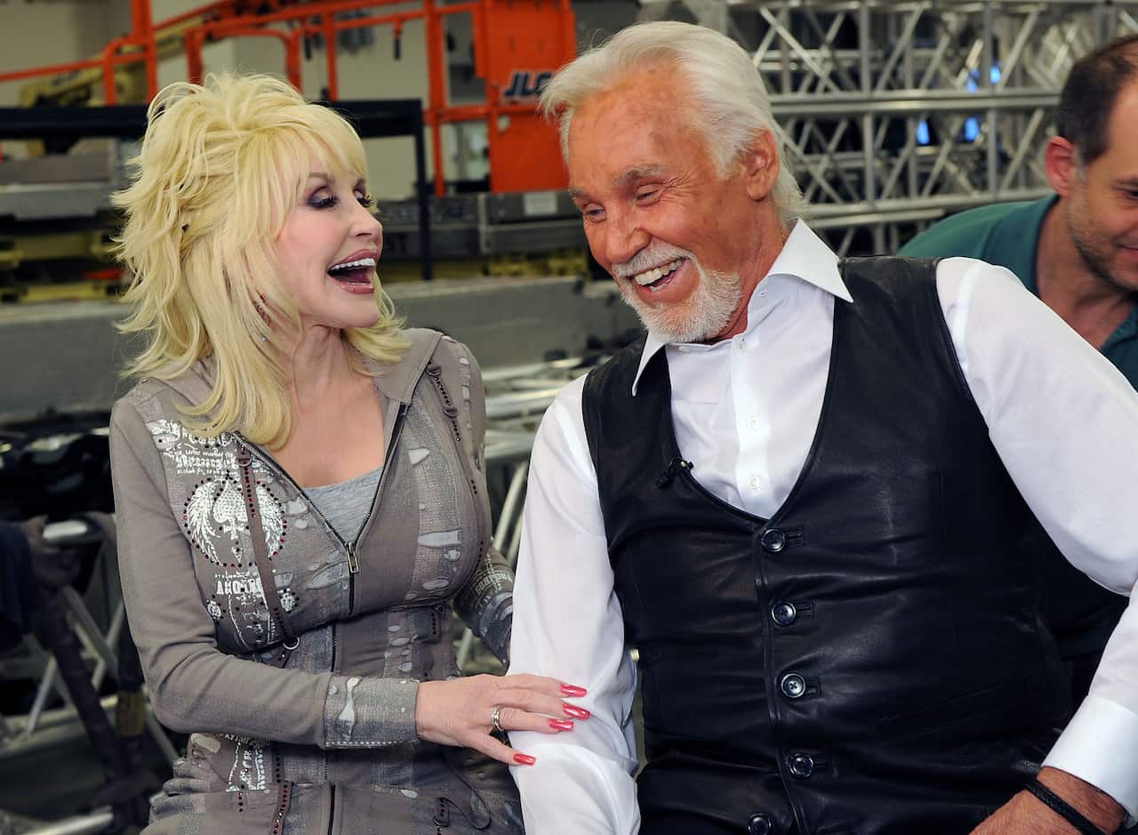 Two people seated side by side in an indoor event or production space, smiling and looking toward each other. One person with light blond hair wears a patterned zip-up top and rests a hand on the other person's arm. The other person wears a white shirt and black leather vest. Metal truss structures and orange equipment are visible in the background.
