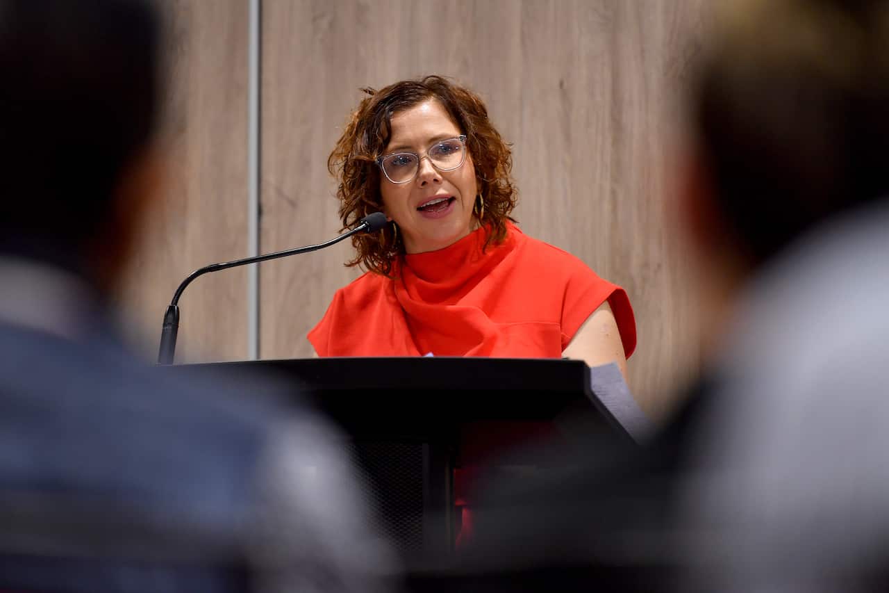 A woman in red top stands in front of a podium and speaks into a microphone.