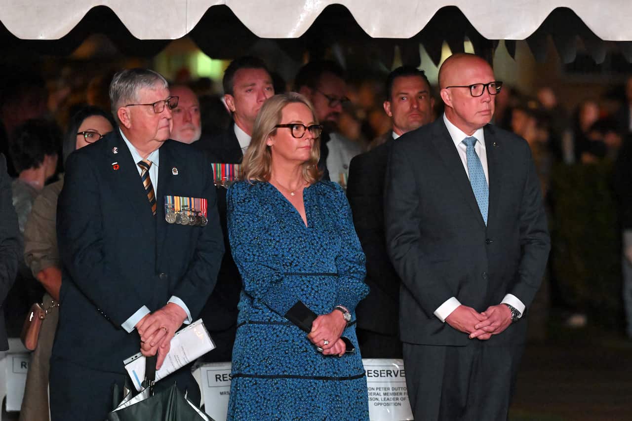 A large crowd of people is standing up. (Left to right) At the front are a man wearing medals, a woman in a blue dress, and a man in a black suit with a blue tie.