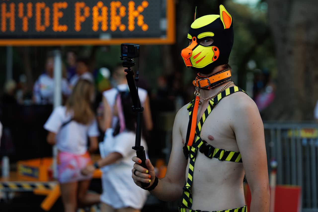 A shirtless person in a black, orange and yellow kinky dog mask holds a selfie stick.