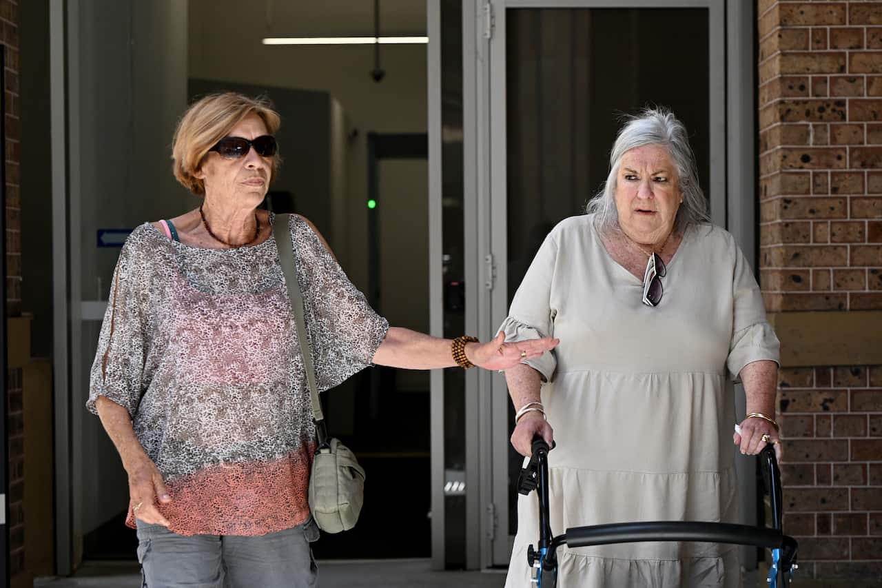 An elderly lady walks using a walker with another elderly lady to her right.